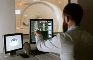 Profesional sanitario analizando im&aacute;genes de una resonancia magn&eacute;tica de columna en una sala de control, con el equipo de RM visible al fondo.